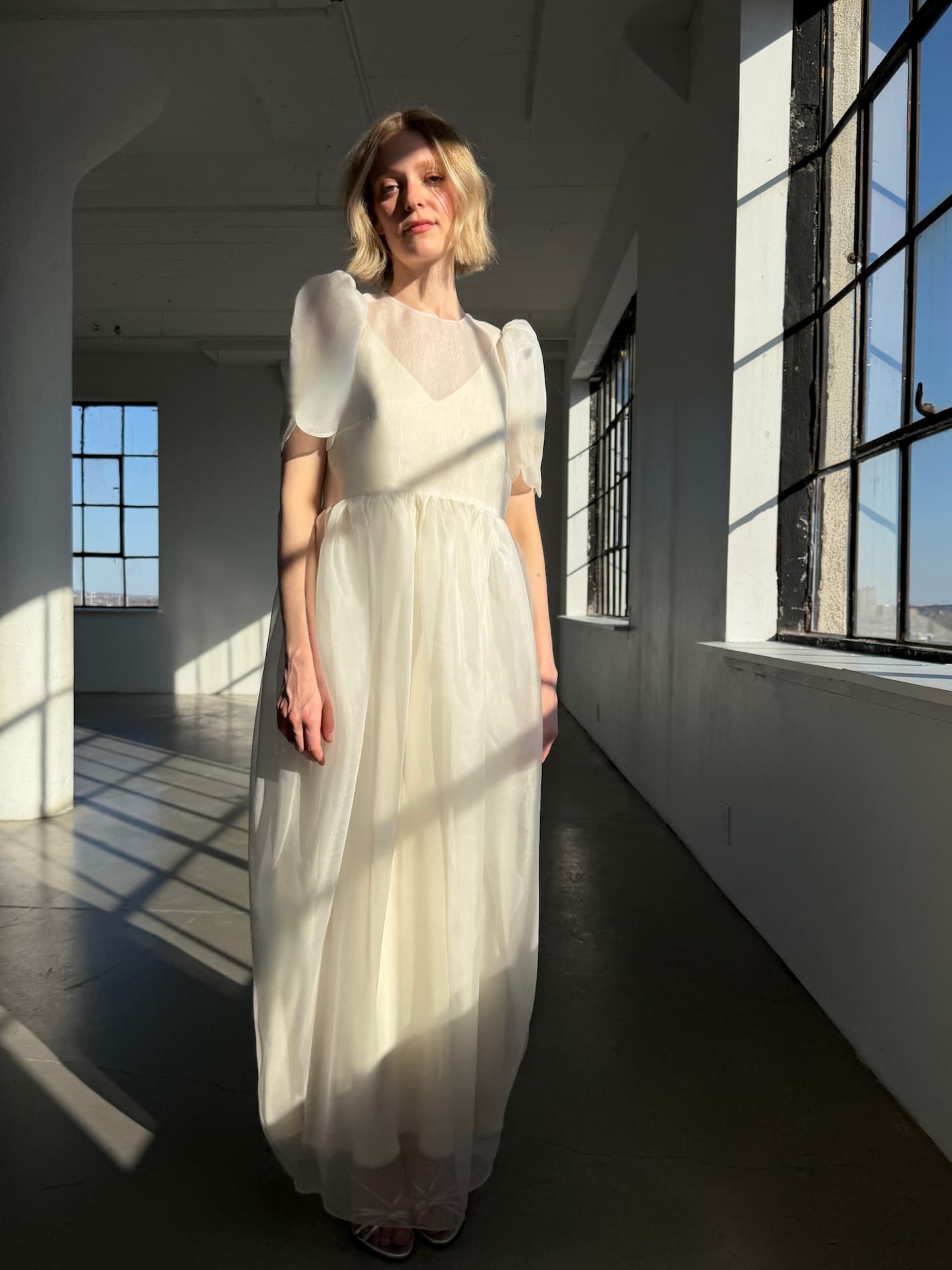 A woman wears the Noémiah Perle Dress — Bridal Edit, standing in a sunlit room with large industrial windows as soft light casts shadows across the floor and her delicate dress.