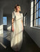 A woman wears the Noémiah Perle Dress — Bridal Edit, standing in a sunlit room with large industrial windows as soft light casts shadows across the floor and her delicate dress.