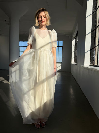 A woman wearing the Perle Dress — Bridal Edit by Noémiah stands in a sunlit room, gently holding the skirt. Sunlight highlights the delicate, sheer fabric and soft puff sleeves of her elegant white dress.