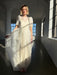 A woman wearing the Perle Dress — Bridal Edit by Noémiah stands in a sunlit room, gently holding the skirt. Sunlight highlights the delicate, sheer fabric and soft puff sleeves of her elegant white dress.