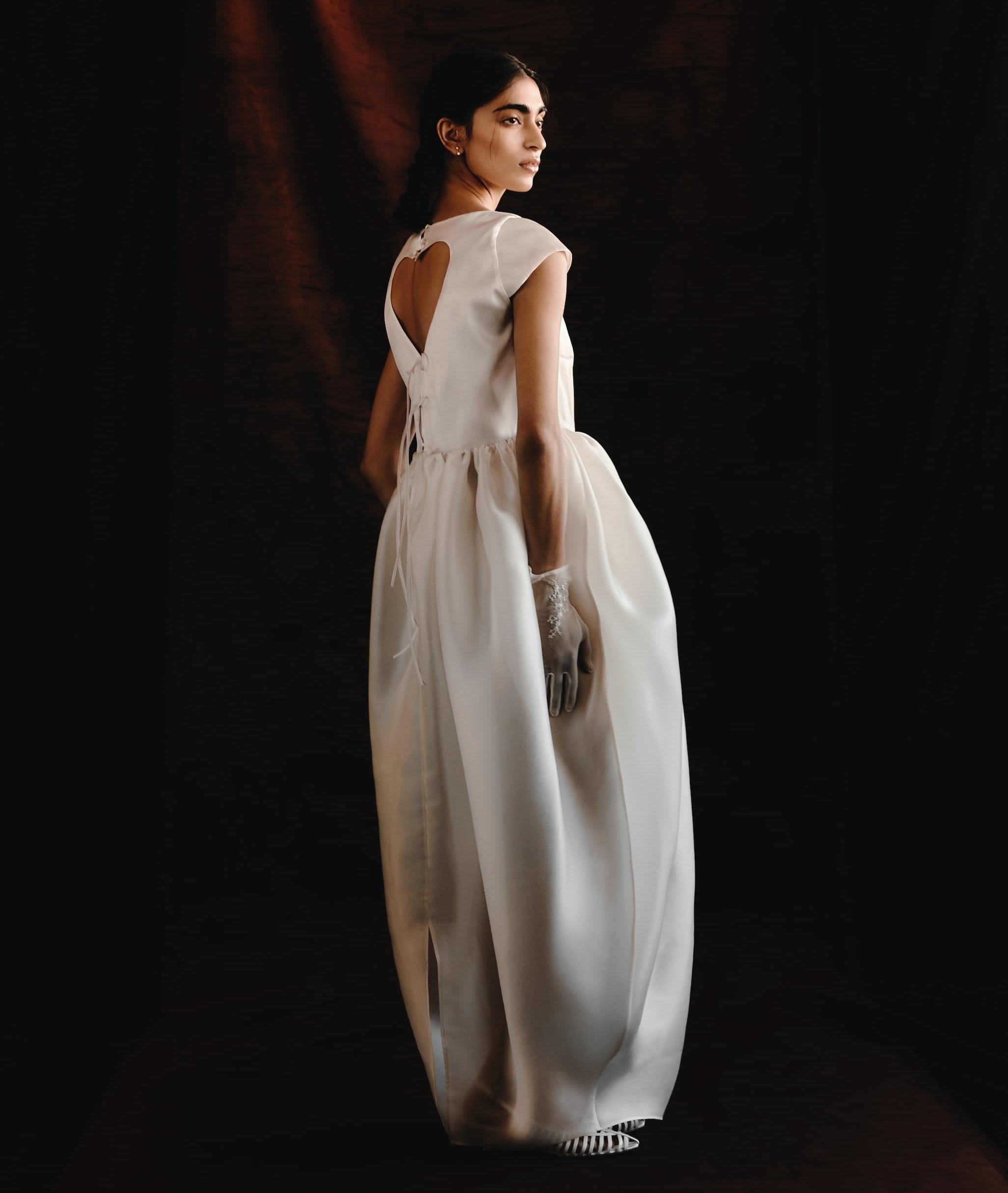A woman in the Noémiah Perle Dress — Bridal Edit, a white silk organza gown with a keyhole back and cap sleeves, stands against a dark backdrop. She wears sheer gloves and her long dark hair is pulled back as she looks over her shoulder.