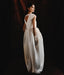 A woman in the Noémiah Perle Dress — Bridal Edit, a white silk organza gown with a keyhole back and cap sleeves, stands against a dark backdrop. She wears sheer gloves and her long dark hair is pulled back as she looks over her shoulder.