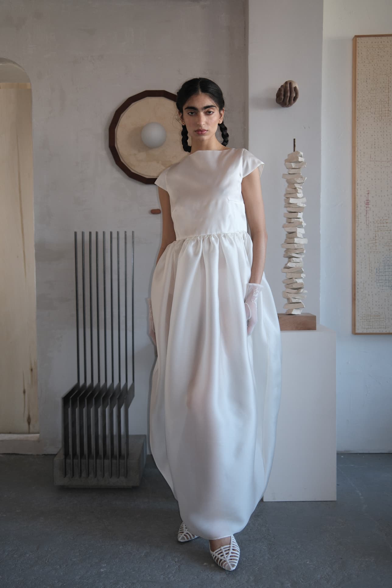 A woman stands indoors on concrete, framed by abstract art and sculpture, wearing Noémiah’s Perle Dress — Bridal Edit: a white, floor-length silk organza gown with cap sleeves and a heart-shaped open back, paired with gloves and white shoes.