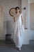 A woman stands indoors on concrete, framed by abstract art and sculpture, wearing Noémiah’s Perle Dress — Bridal Edit: a white, floor-length silk organza gown with cap sleeves and a heart-shaped open back, paired with gloves and white shoes.