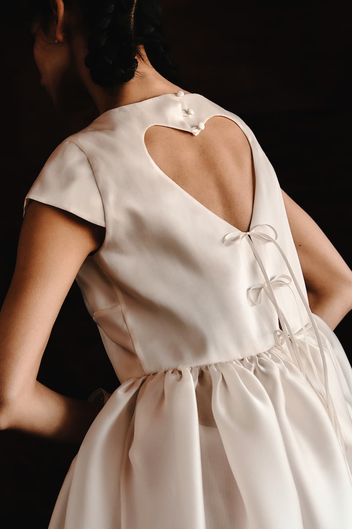 A woman wears the Perle Dress — Bridal Edit by Noémiah, a silk organza floor-length gown with a heart-shaped cutout, decorative back buttons, ribbon ties, and cap sleeves. A dark background highlights its elegant details.
