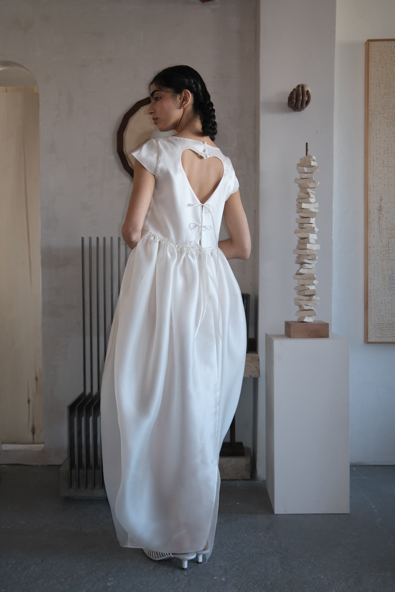 A woman stands indoors, facing away, wearing the Perle Dress — Bridal Edit by Noémiah. The silk organza gown features a heart-shaped open back, flowing floor-length skirt, and back ties. Modern art and decor appear in the background.