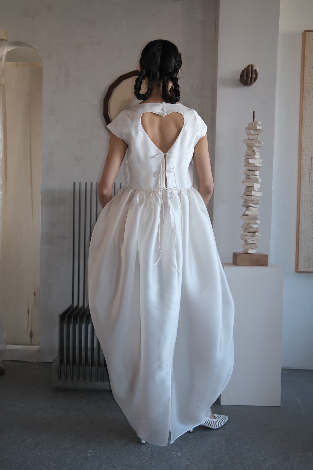 A woman in the Noémiah Perle Dress — Bridal Edit, a silk organza gown with a heart-shaped open back, stands indoors facing away. The full-skirted, floor-length dress ties at the back; the room is minimally decorated in neutral tones.