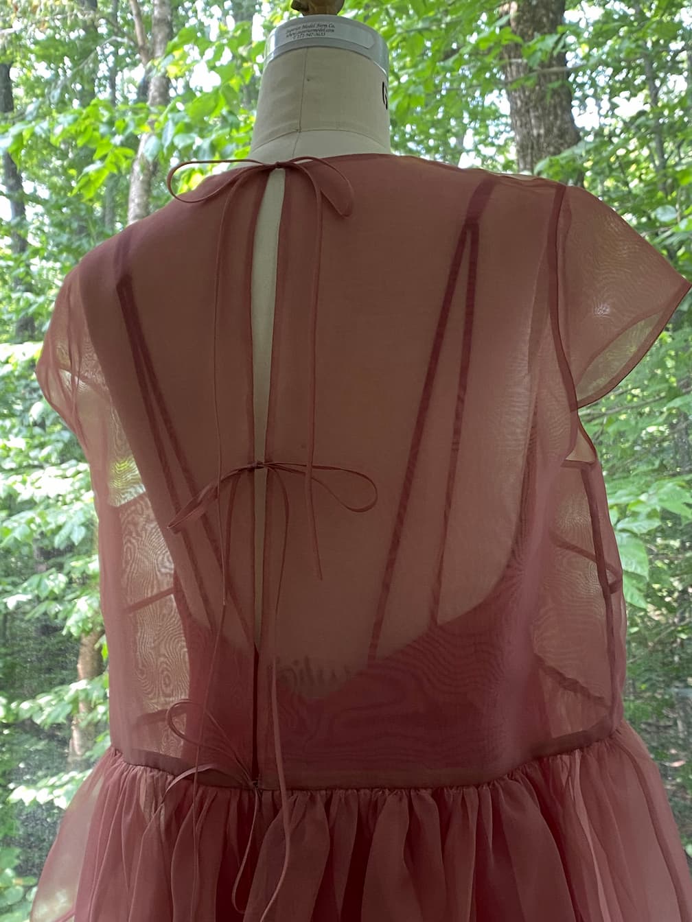 The Perle Dress — Classic by Noémiah, a sheer rose silk organza piece with an open back and two thin ties, is displayed on a dress form before a window framing green trees.