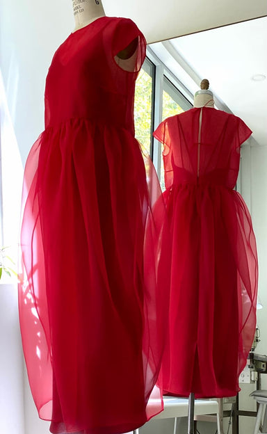 The Perle Dress — Classic by Noémiah is a sheer, bright red silk organza dress with short sleeves, a fitted bodice, and voluminous skirt. Shown on a mannequin before a mirror, it displays both the elegant front and back views for an ethereal effect.