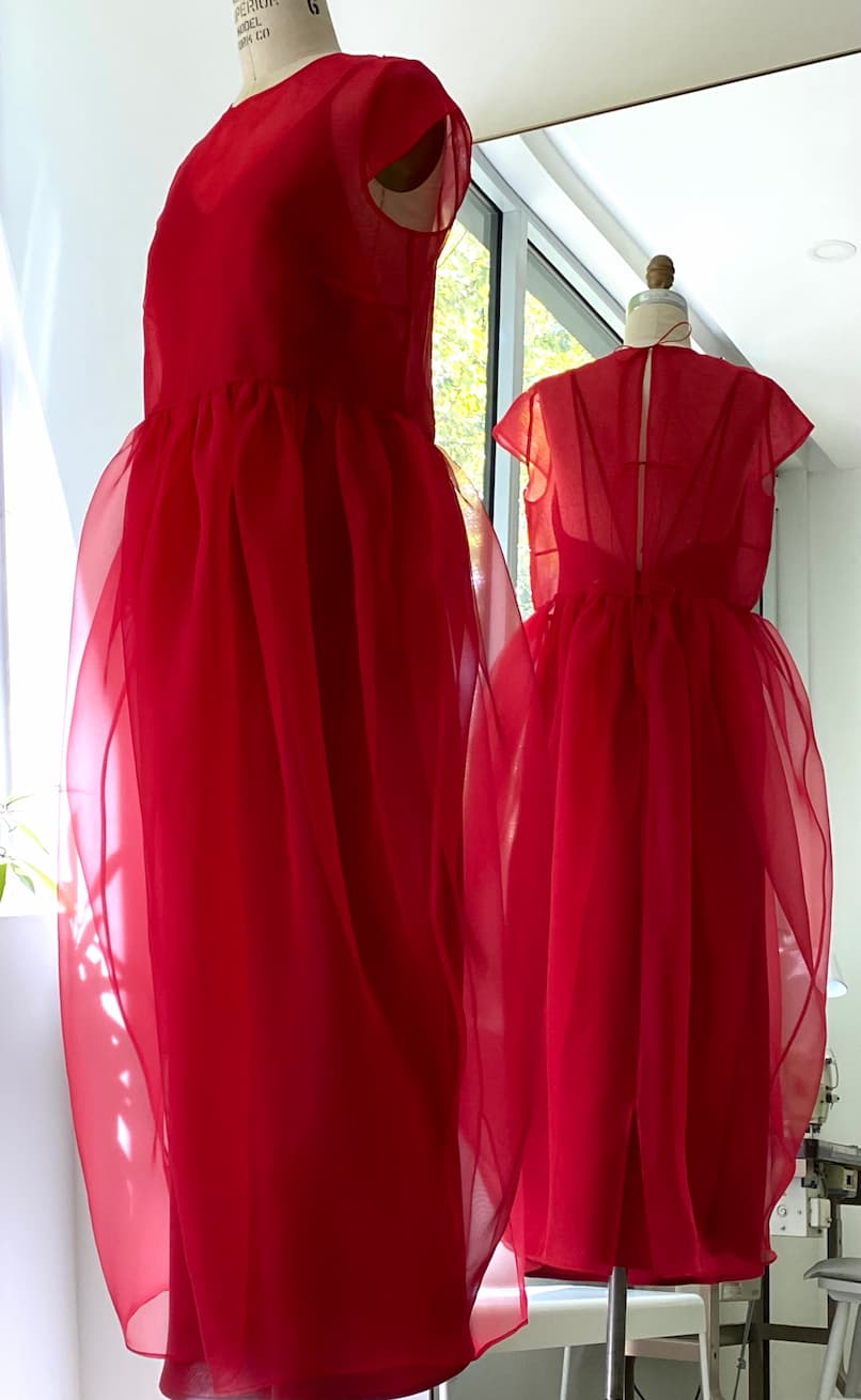 The Perle Dress — Classic by Noémiah is a sheer, bright red silk organza dress with short sleeves, a fitted bodice, and voluminous skirt. Shown on a mannequin before a mirror, it displays both the elegant front and back views for an ethereal effect.