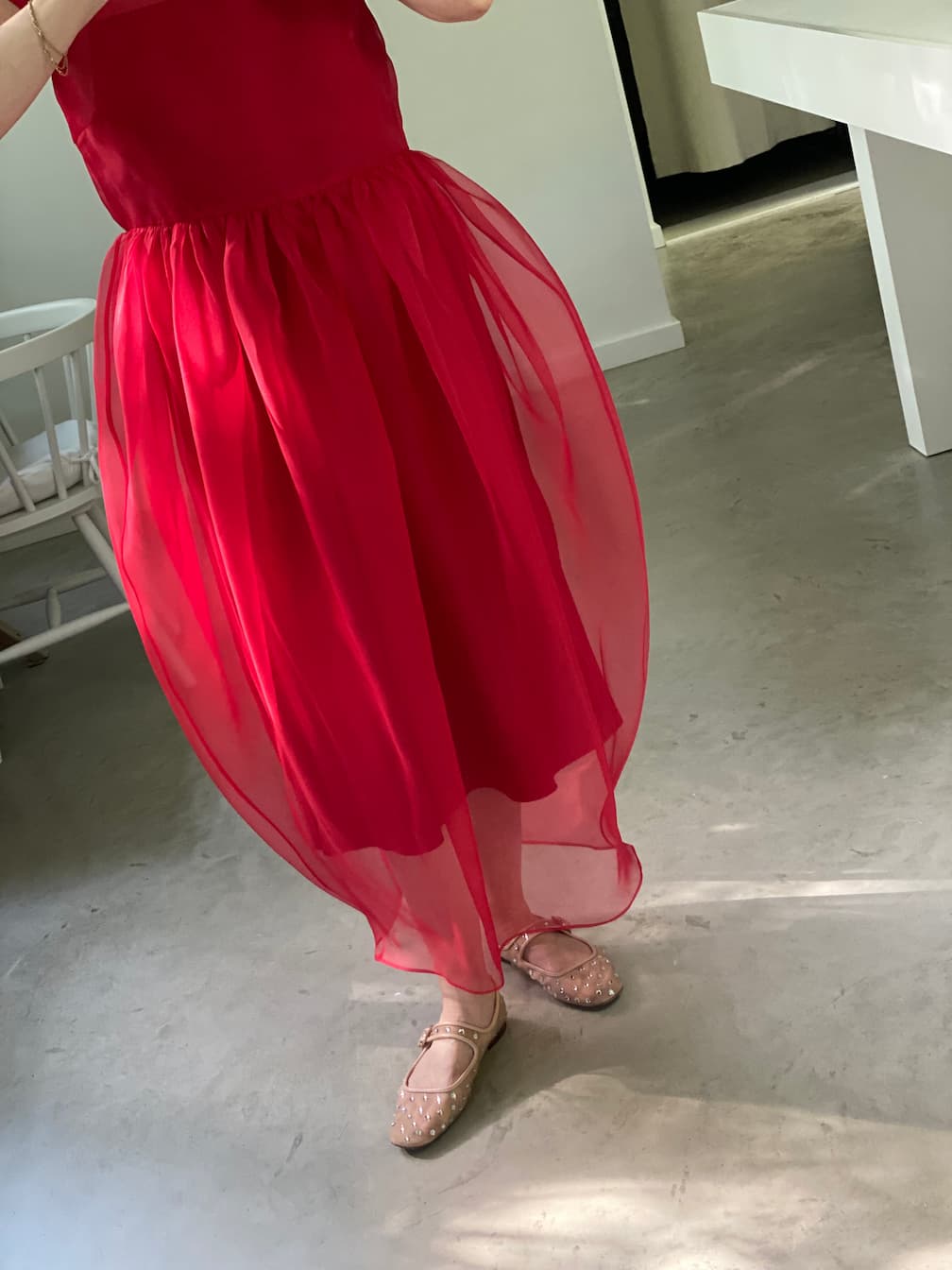 Wearing the Noémiah Perle Dress — Classic, a bright pink sheer design with a delicate overlay, this person stands indoors on a polished floor in light pink ballet flats. The cropped frame highlights an ethereal silhouette.