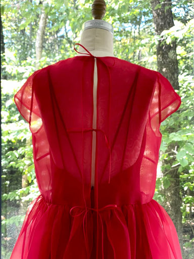 The Perle Dress — Classic by Noémiah, a red silk organza dress with two thin back ties and soft pleats, is displayed on a dress form. Sunlight through green trees highlights its ethereal silhouette.