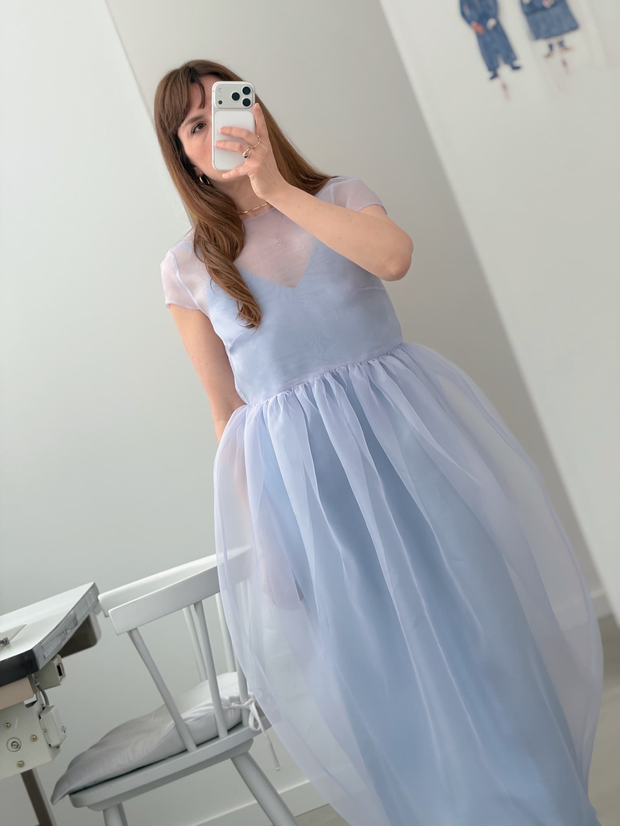 A woman wears the Noémiah Perle Dress — Classic, a light purple sheer piece, while taking a mirror selfie in a minimalistic room with white walls, artwork, and a white chair in the background.