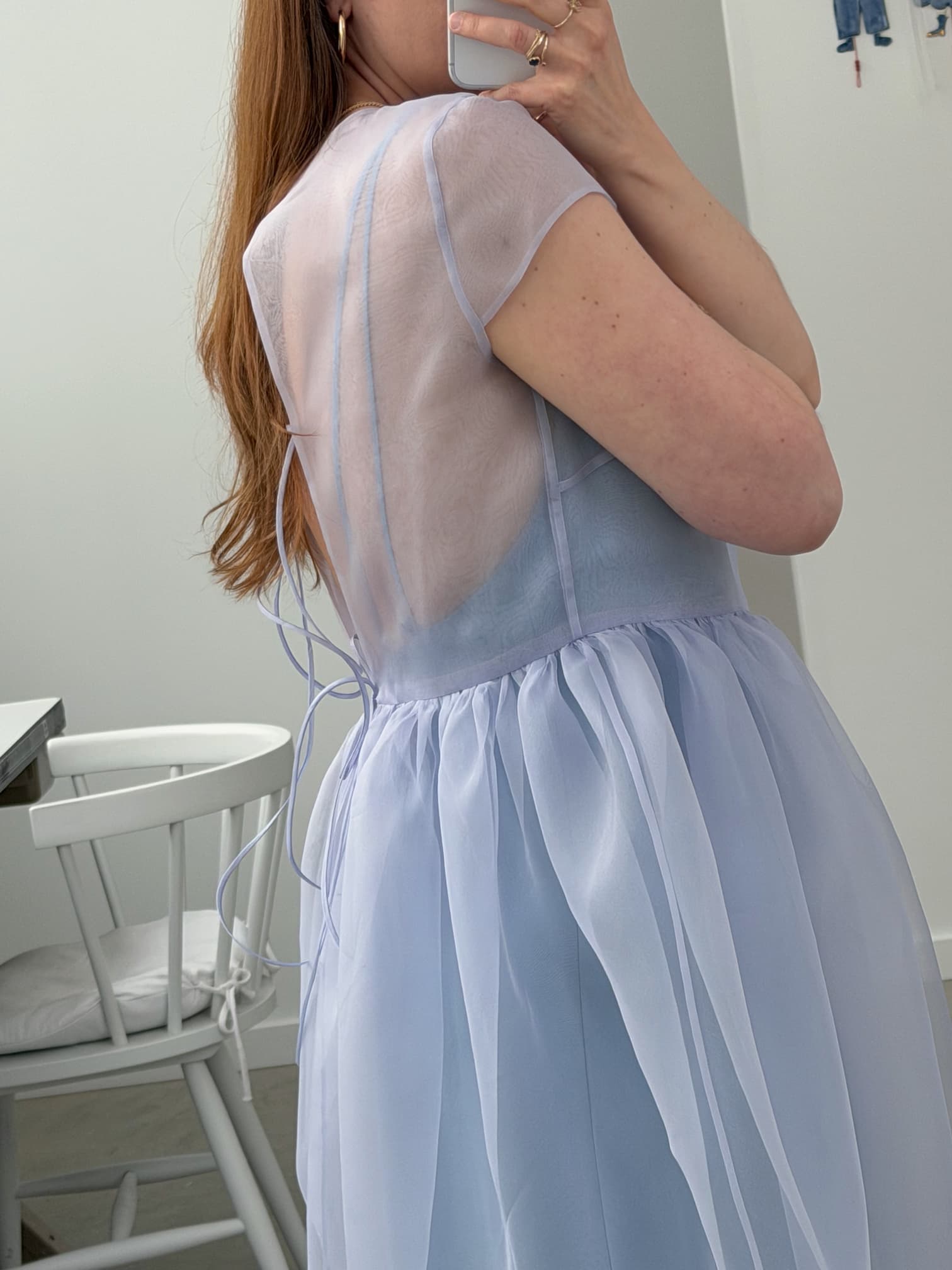 A person taking a mirror selfie shows off the back of the Perle Dress — Classic by Noémiah in light blue silk organza with delicate ribbon ties. The sheer upper part flows into a soft skirt, and a white wooden chair appears in the background.