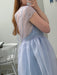 A person taking a mirror selfie shows off the back of the Perle Dress — Classic by Noémiah in light blue silk organza with delicate ribbon ties. The sheer upper part flows into a soft skirt, and a white wooden chair appears in the background.
