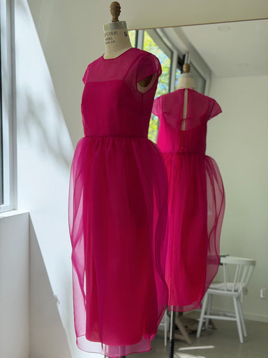 The Noémiah Perle Dress — Classic, a vibrant fuchsia pink with sheer silk organza short sleeves and overlay skirt, is displayed on a dress form in a bright room, its ethereal silhouette reflected in the mirror.