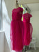 The Noémiah Perle Dress — Classic, a vibrant fuchsia pink with sheer silk organza short sleeves and overlay skirt, is displayed on a dress form in a bright room, its ethereal silhouette reflected in the mirror.