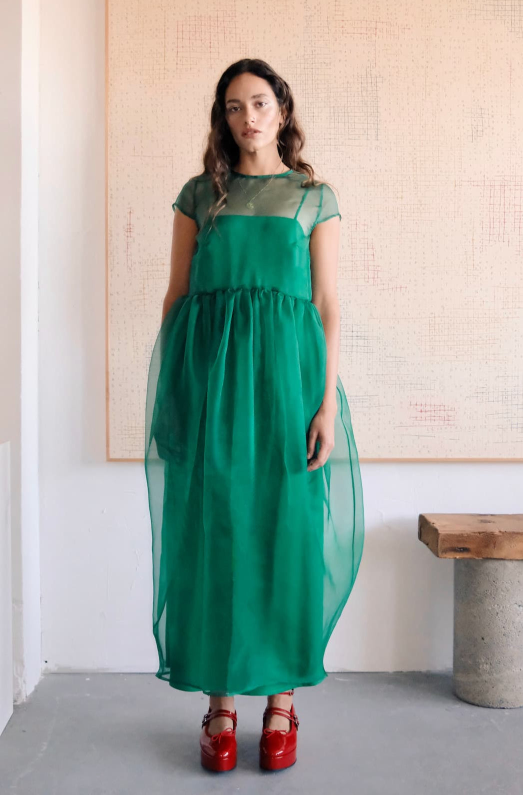 A woman stands indoors wearing the Perle Dress — Classic by Noémiah, a green sheer silk organza dress with short sleeves and a gathered waist over a matching slip. She completes the look with shiny red platform shoes. An abstract painting hangs behind her.