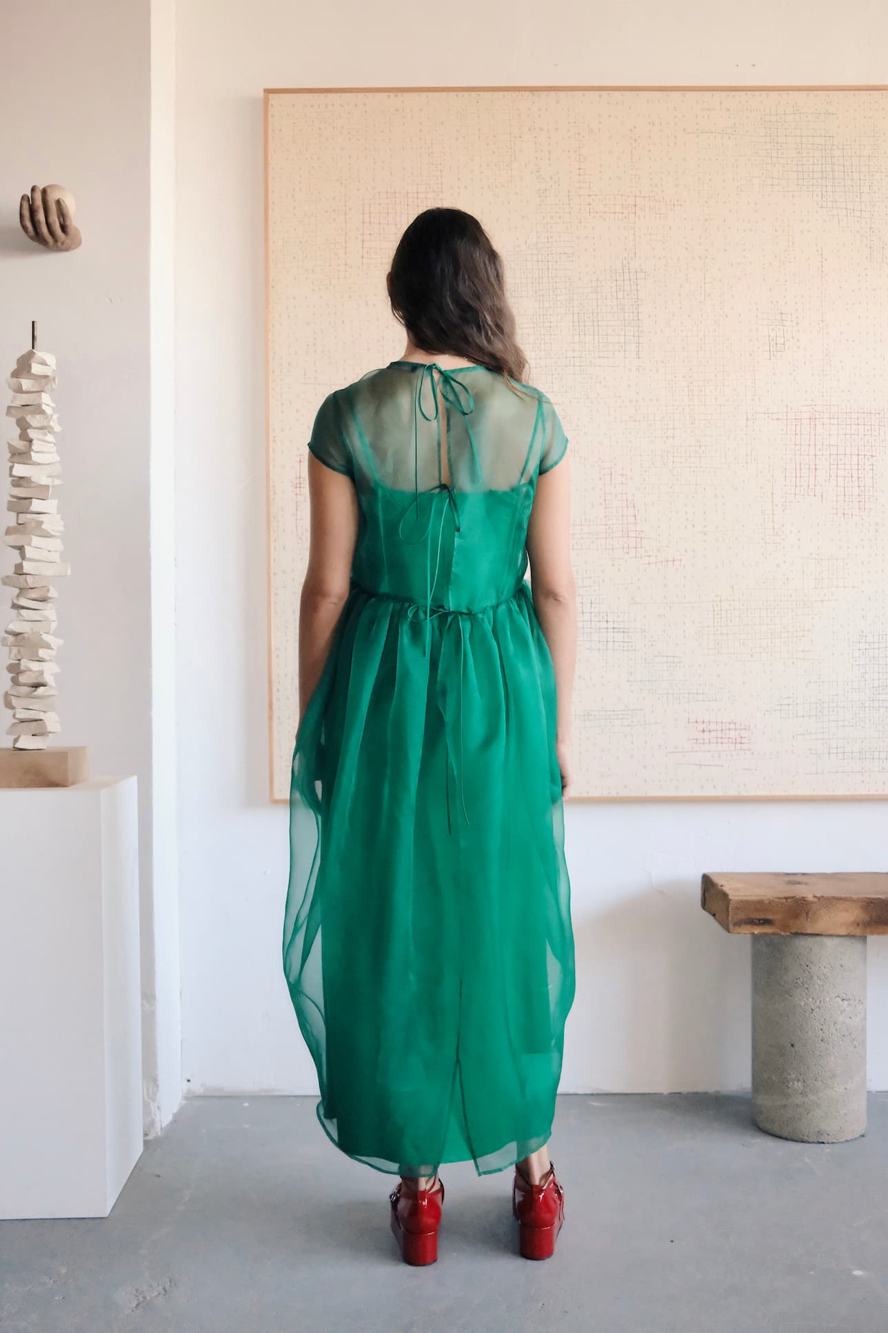 A person faces a large canvas, wearing the Perle Dress by Noémiah and red platform heels. The minimally decorated room features a sculpture on a white pedestal and a wooden bench, with white walls creating an ethereal silhouette and elegant atmosphere.