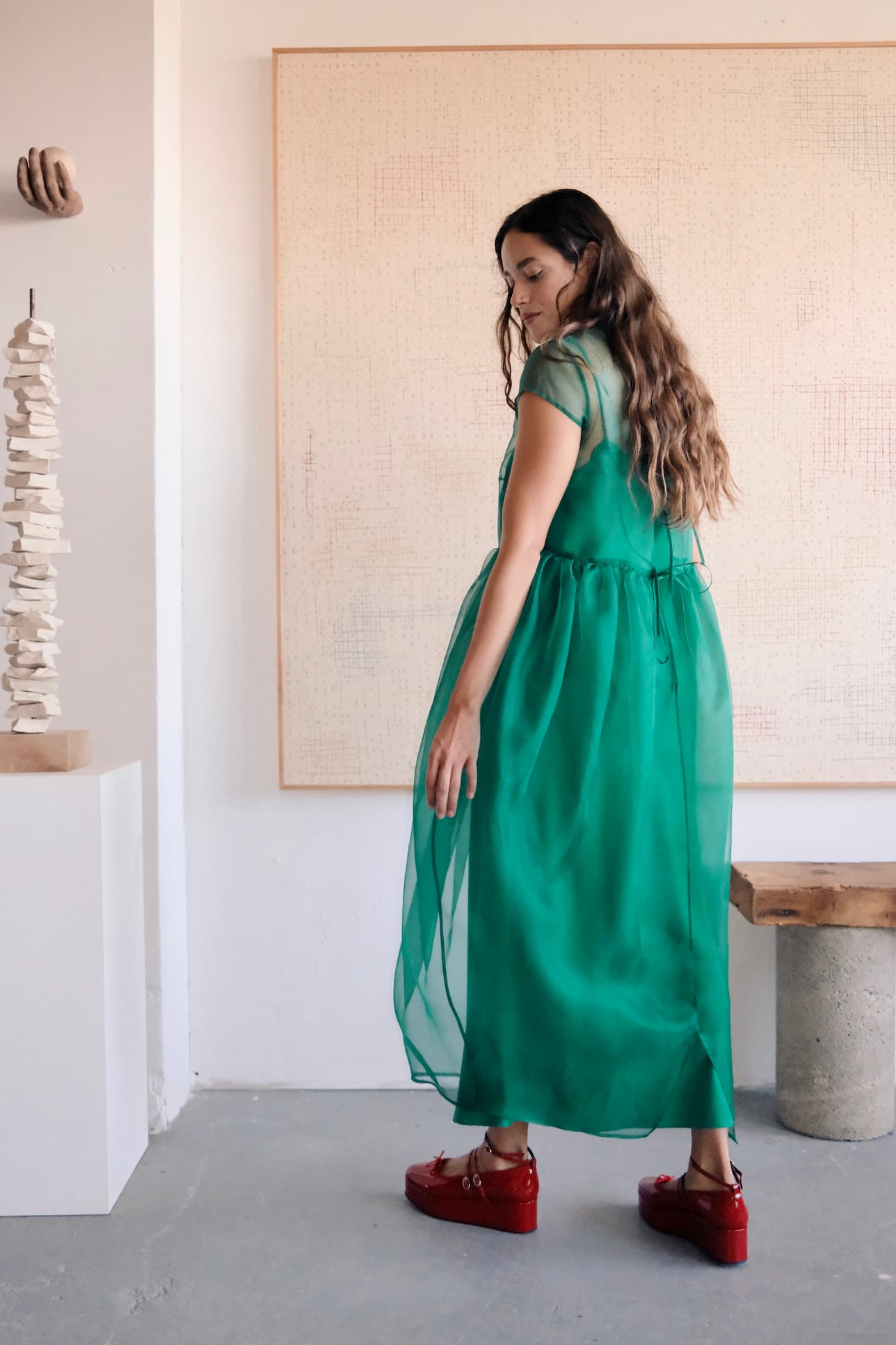 In an art gallery, a woman with an ethereal silhouette stands near a minimalist sculpture and large abstract canvas. Her long hair flows over Noémiahs sheer green Perle Dress in silk organza, while red platform shoes add a bold touch to her ensemble.