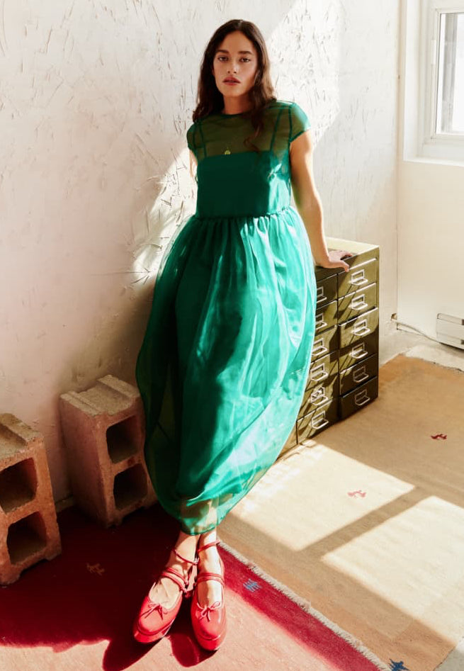 A woman in the Perle Dress by Noémiah and red shoes leans against a wall in a sunlit room with cinder blocks, green boxes, and colorful rugs, casting an ethereal silhouette.