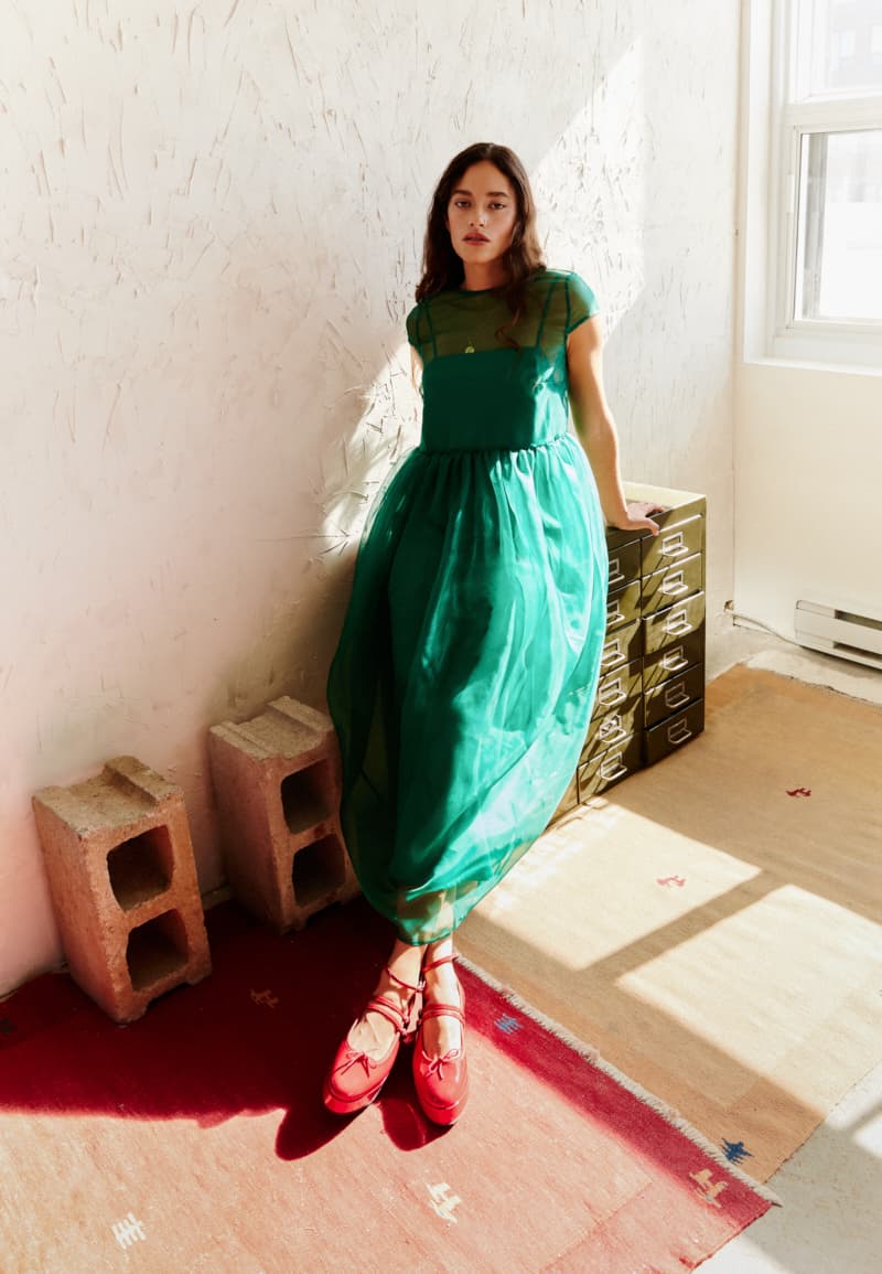 A woman in the Perle Dress by Noémiah and red shoes leans against a wall in a sunlit room with cinder blocks, green boxes, and colorful rugs, casting an ethereal silhouette.