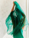 A person in the Noémiah Perle Dress, made of flowing green silk organza, raises their arms partially covering their face, illuminated by soft light against a textured white wall.