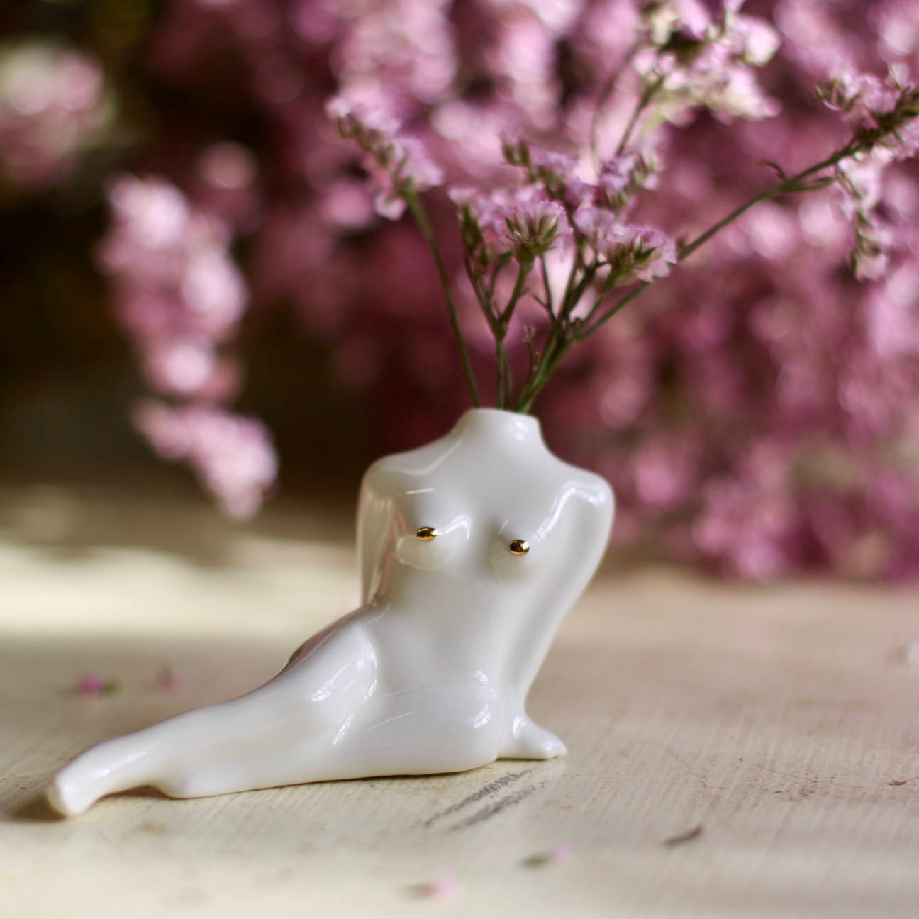 A Porcelain Bud Vase by Noémiah, shaped like a reclining human torso, cradles delicate pink flowers. It is set on a wooden surface, enhancing your space with its handcrafted charm, as additional pink flowers create a gentle blur in the background.