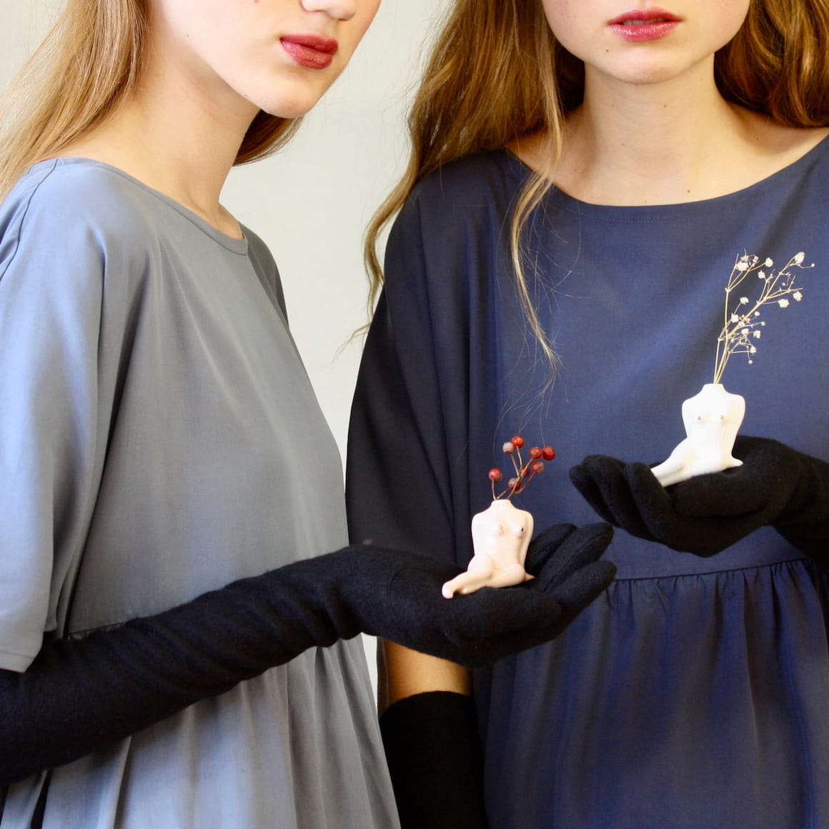 Dressed in grey dresses and black gloves, two individuals hold small porcelain bud vases from Noémiah. The vases, abstract in design with branch-like features resembling torsos, contribute to a minimalist and artistic scene that blends fashion with art. Each vase complements a self-care ritual like a unique handmade decoration.