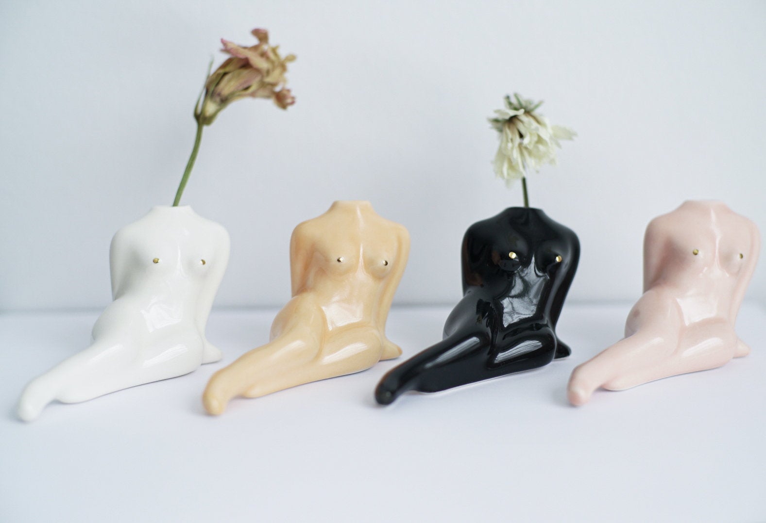 The Porcelain Bud Vase by Noémiah, available in white, beige, black, and pink, adds a handmade decorative touch with its reclining female torso shape. Each vase includes a small hole perfect for displaying flower stems, beautifully complemented by two dried flowers in the white and black versions.