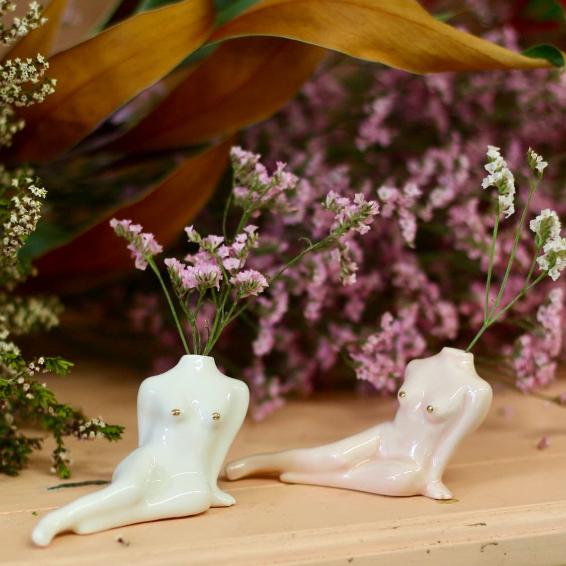 A pair of Porcelain Bud Vases from Noémiah, shaped like reclining human torsos with flowering plants sprouting from them, set a serene scene against a lush floral and verdant backdrop. These handmade decorations boast a glossy finish, one in white and the other in pastel pink, making them ideal for any self-care ritual.