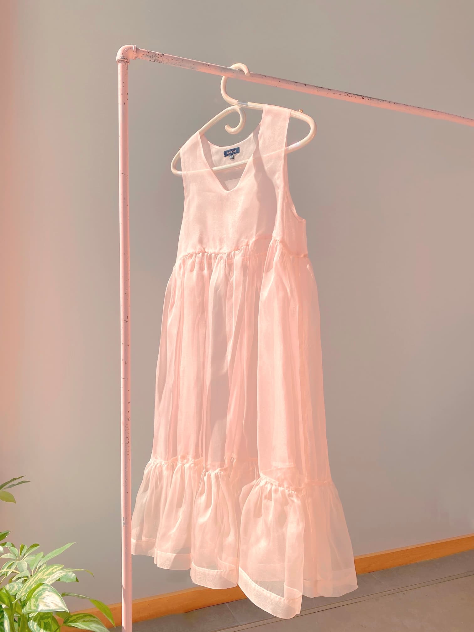 A Porcelain Hanger by Noémiah displays a delicate pink sheer dress with an iridescent luster on a light pink metal rack against a soft gray wall. Sunlight casts gentle shadows, highlighting the fabric's texture, while a small green plant in the bottom left corner adds a touch of nature to this serene setting.