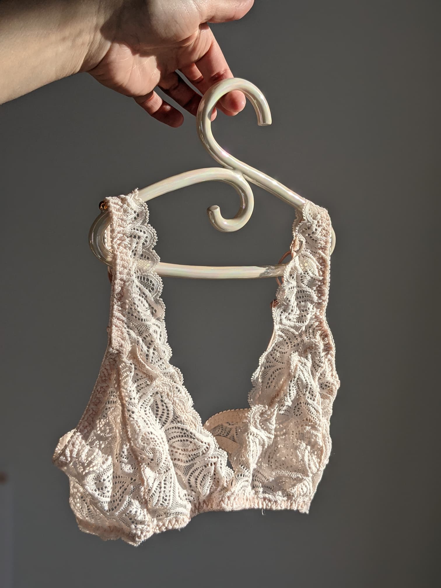 A hand holds a Porcelain Hanger by Noémiah, displaying a delicate cream-colored lace bralette against a neutral background. The bralette, akin to wedding dresses, showcases intricate lace patterns and soft fabric, with sunlight emphasizing its iridescent luster.