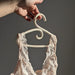A hand holds a Porcelain Hanger by Noémiah, displaying a delicate cream-colored lace bralette against a neutral background. The bralette, akin to wedding dresses, showcases intricate lace patterns and soft fabric, with sunlight emphasizing its iridescent luster.