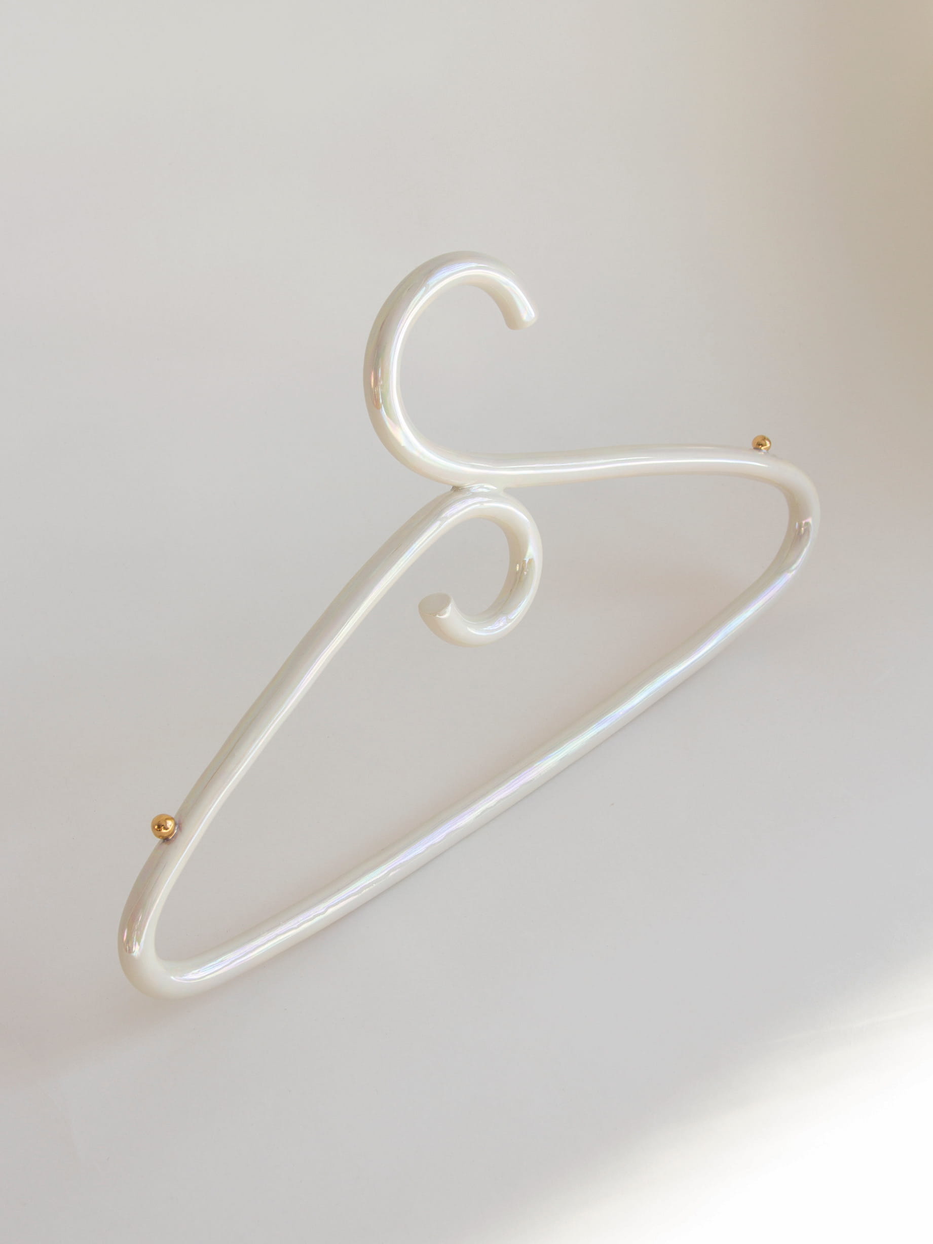 The Porcelain Hanger by Noémiah is a sleek, modern coat hanger with an iridescent luster and elegant curves, highlighted by gold accents at the joints. Its glossy white finish stands out beautifully against a soft, light background, accentuating its minimalist and stylish design.