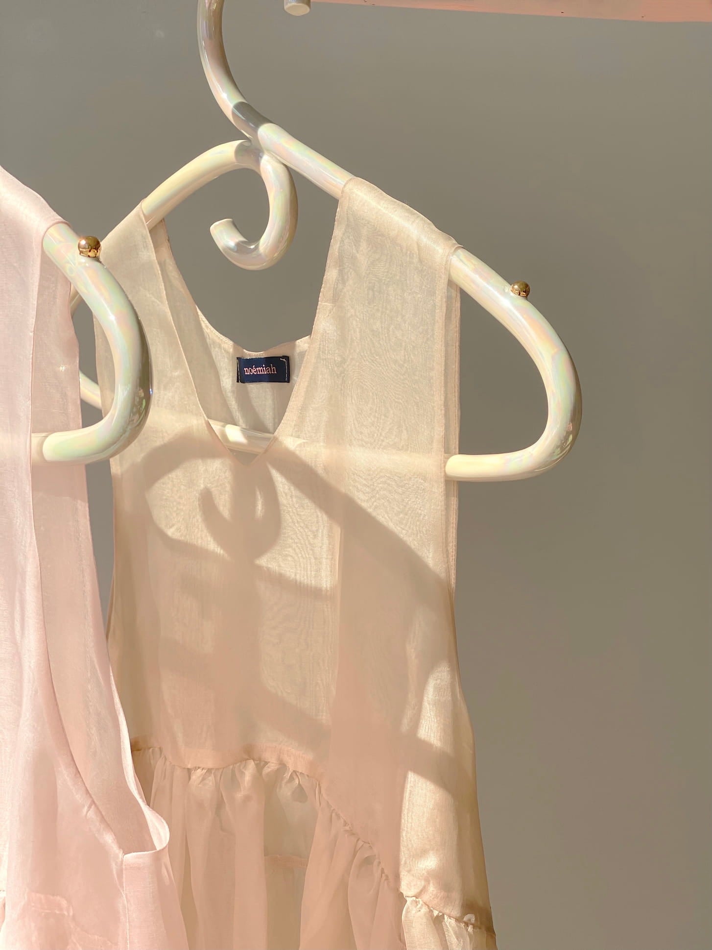 A delicate, translucent white dress with an iridescent luster hangs on a Porcelain Hanger from Noémiah. Sunlight casts soft shadows, creating a serene and elegant ambiance. The muted gray background highlights the dress's light and airy fabric, reminiscent of ethereal wedding dresses.