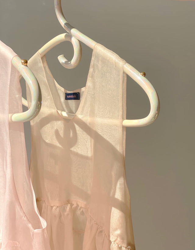 A delicate, translucent white dress with an iridescent luster hangs on a Porcelain Hanger from Noémiah. Sunlight casts soft shadows, creating a serene and elegant ambiance. The muted gray background highlights the dress's light and airy fabric, reminiscent of ethereal wedding dresses.