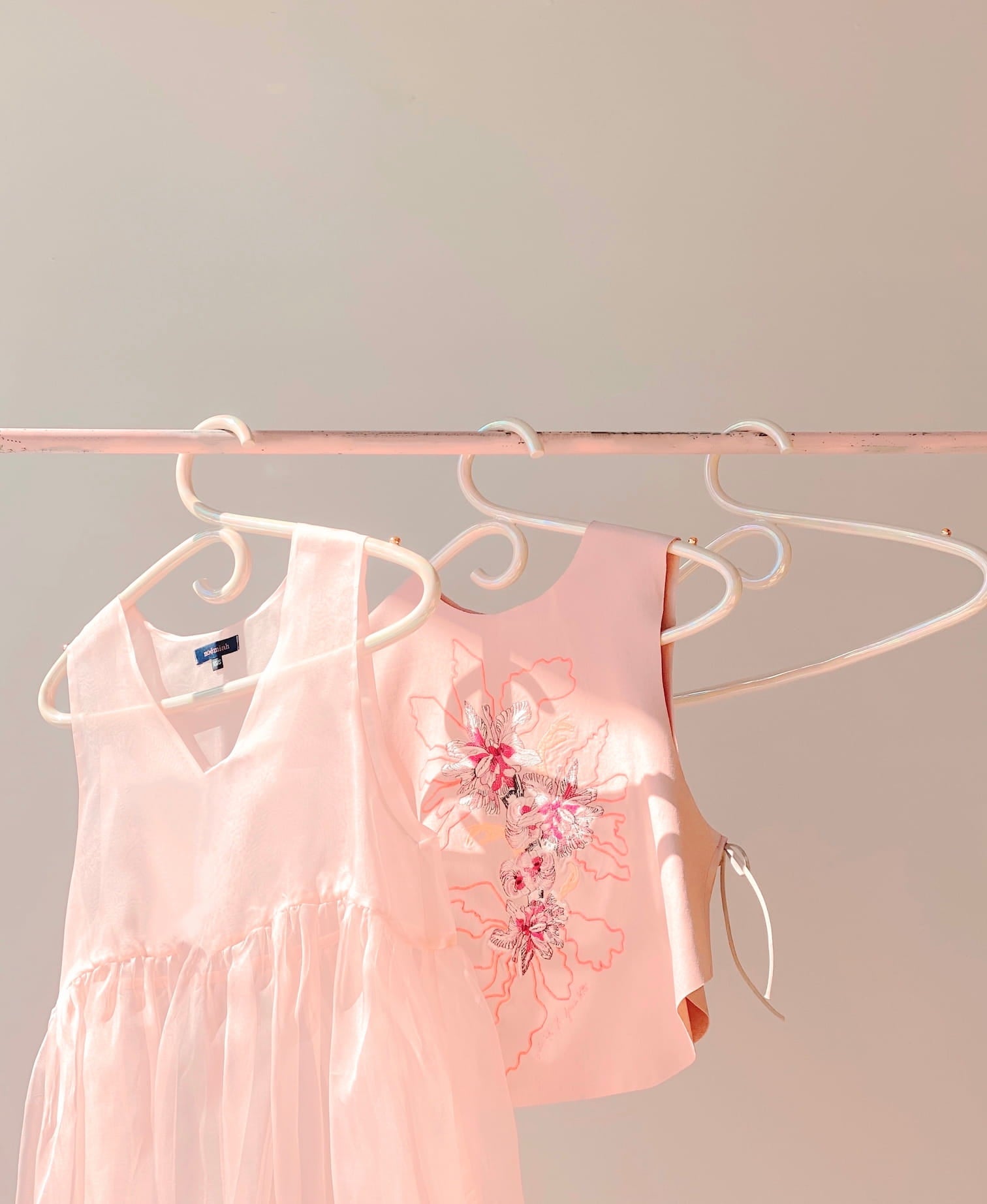 Two light pink dresses hang on Noémiah's Porcelain Hanger against a neutral background. One dress shimmers with an iridescent luster, featuring a sheer skirt. The other is sleeveless with embroidered floral designs that evoke the delicate beauty of handcrafted porcelain.