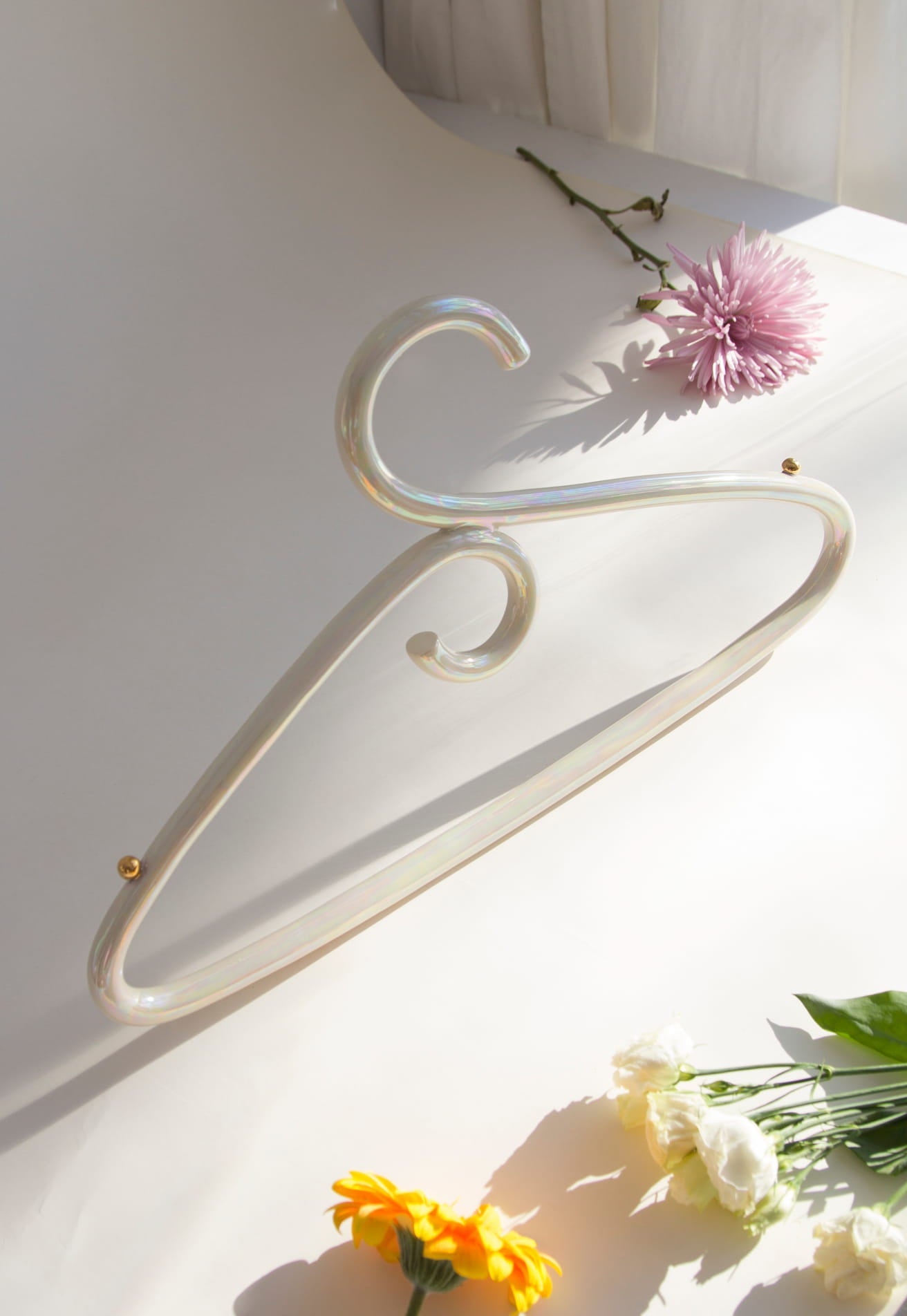 A Porcelain Hanger by Noémiah stands upright against a cream backdrop with soft lighting, its iridescent luster gently catching the eye. Surrounding it are fresh flowers: a pink chrysanthemum, a yellow marigold, and white roses, adding a touch of color and elegance reminiscent of handcrafted porcelain.