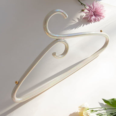 A Porcelain Hanger by Noémiah stands upright against a cream backdrop with soft lighting, its iridescent luster gently catching the eye. Surrounding it are fresh flowers: a pink chrysanthemum, a yellow marigold, and white roses, adding a touch of color and elegance reminiscent of handcrafted porcelain.