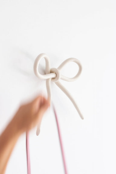 A hand holding pink cables reaches towards the Porcelain Hook by Noémiah, designed like a bow, cleverly utilizing wall space against the plain white backdrop.