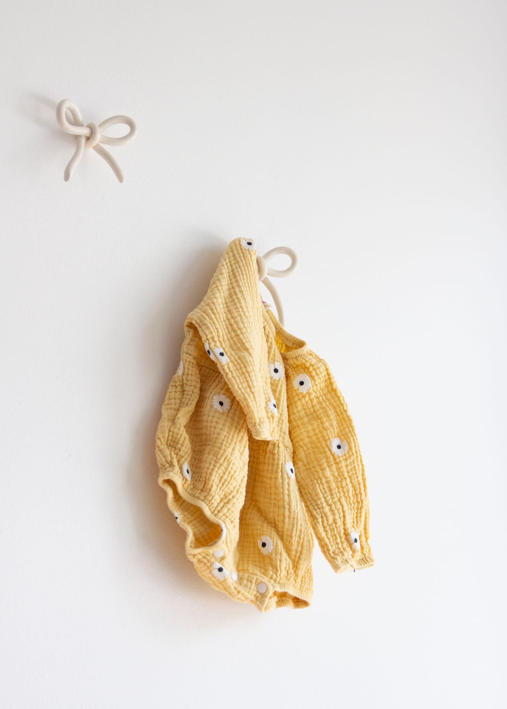 A yellow children's onesie with a textured pattern and small embroidered faces hangs on a white wall from Noémiah's Porcelain Hook. Another similar hook is visible above it, adding charm to the wall space.