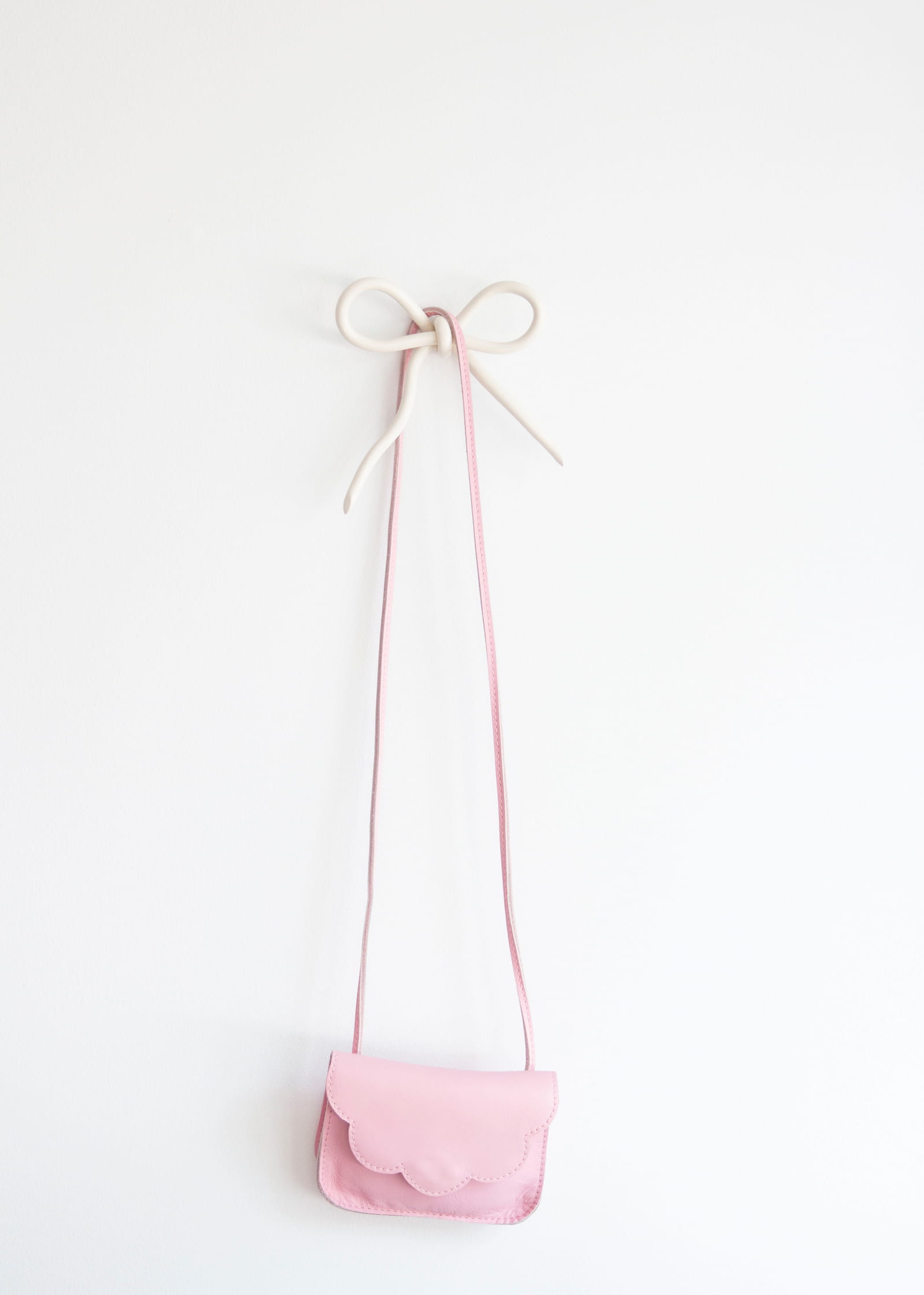 A beautifully handcrafted small pink purse with a scalloped flap elegantly hangs from Noémiah's Porcelain Hook on a white wall. The strap is artfully tied into a bow at the top, securely suspended from the delicate hook.