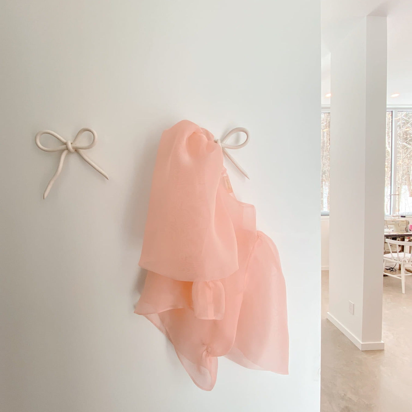 A light pink, sheer scarf hangs elegantly on a white wall from a small, bow-shaped Porcelain Hook by Noémiah. The bright, minimalistic room features white walls and a concrete floor, with sunlight streaming in through large windows that highlights the carefully curated wall space.