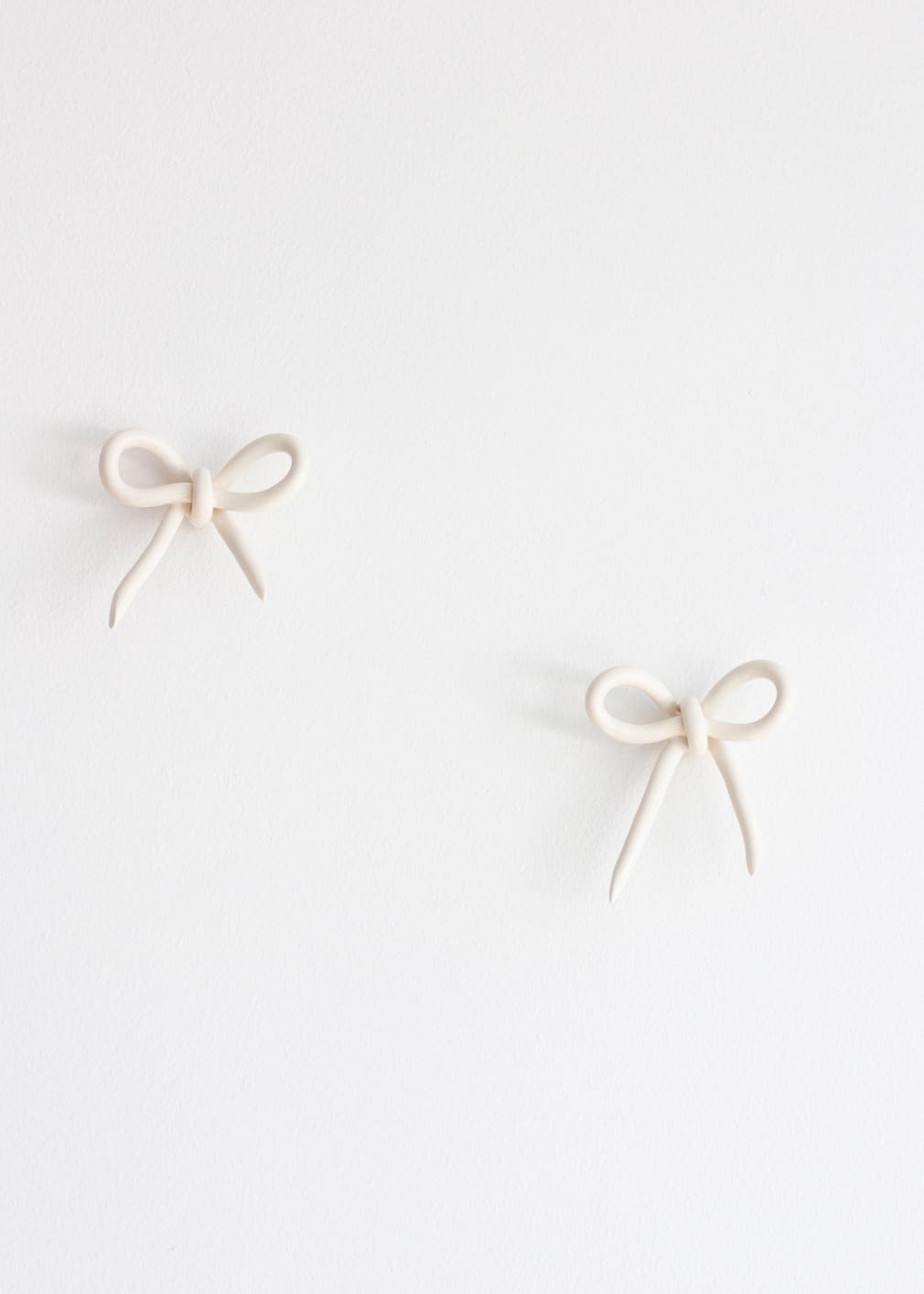 Two small, bow-shaped Porcelain Hooks by Noémiah, featuring a handcrafted design in beige, are mounted on a plain white wall.