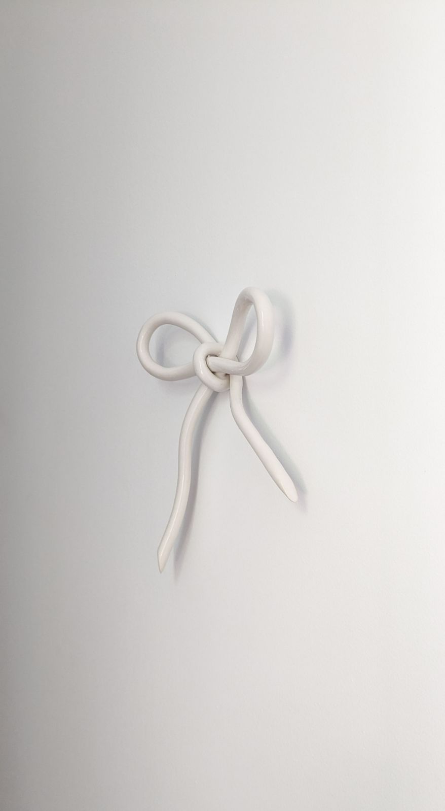 A minimalist photo features the Porcelain Hook by Noémiah, a white ceramic piece with a graceful, loosely tied bow design and long flowing ends. Its handcrafted elegance enhances the wall space as it is mounted on a plain white surface.