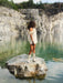 A person stands on a large rock wearing the Rosie Dress, a cream silk taffeta mini dress by Noémiah, paired with boots. They are beside a calm lake with clear, green water, surrounded by rocky cliffs and dense trees in the background.