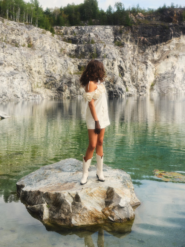 A person stands on a large rock wearing the Rosie Dress, a cream silk taffeta mini dress by Noémiah, paired with boots. They are beside a calm lake with clear, green water, surrounded by rocky cliffs and dense trees in the background.