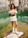 A person wearing the Rosie Dress by Noémiah, a silk taffeta mini dress with floral details, paired with white cowboy boots, stands confidently on a rocky surface beside a body of green water, set against a backdrop of rugged stone cliffs.