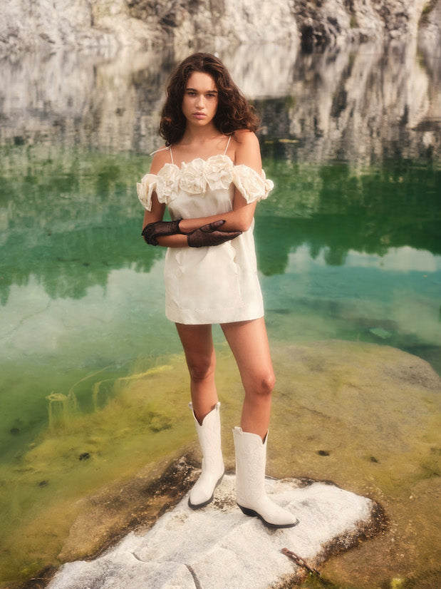 A person wearing the Rosie Dress by Noémiah, a silk taffeta mini dress with floral details, paired with white cowboy boots, stands confidently on a rocky surface beside a body of green water, set against a backdrop of rugged stone cliffs.